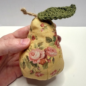 May include: A decorative pear-shaped fabric item with a yellow floral pattern featuring pink roses and green leaves. The pear has a green crocheted leaf and a twine stem, creating a charming, handcrafted aesthetic.