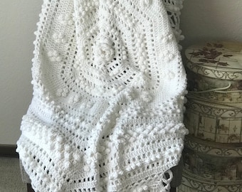May include: A white, handmade crochet blanket draped over a chair. The blanket features a detailed floral and textured design with bobbles and a scalloped edge. The blanket is made of soft, white yarn and is a cozy home decor item.