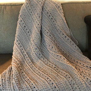 May include: A light gray, hand-crocheted blanket with a textured, diagonal pattern. The blanket is draped over a gray sofa, showcasing its soft, plush appearance. The crochet stitches create a visually appealing, cozy texture.