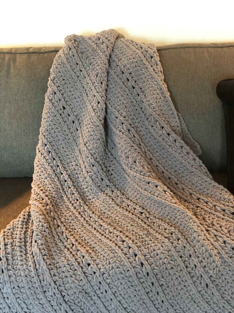 May include: A light gray, hand-crocheted blanket with a textured, diagonal pattern. The blanket is draped over a gray sofa, showcasing its soft, plush appearance. The crochet stitches create a visually appealing, cozy texture.