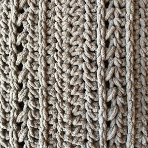 May include: Close-up of a soft, chunky knit blanket in a muted gray color. The blanket features a textured pattern with vertical columns and a woven design, creating a cozy and inviting look. The thick yarn gives it a plush appearance.