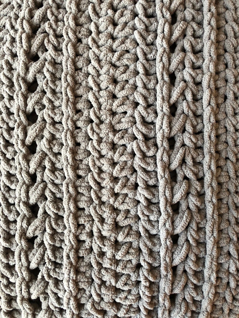 May include: Close-up of a soft, chunky knit blanket in a muted gray color. The blanket features a textured pattern with vertical columns and a woven design, creating a cozy and inviting look. The thick yarn gives it a plush appearance.