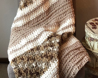 May include: A handmade, chunky knit blanket in shades of cream, brown, and tan, draped over a gray chair. The blanket features a textured, diagonal pattern with alternating stripes and a detailed stitch design. The cozy throw is perfect for home decor.