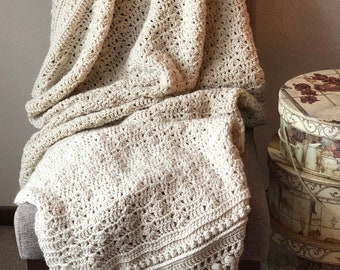 May include: A cream-colored, hand-crocheted blanket with a textured pattern and decorative pom-poms. The blanket is draped over a gray chair. The intricate design and soft color palette create a cozy, inviting aesthetic.