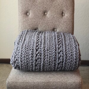 May include: A gray, chunky knit blanket rests on a button-tufted upholstered chair. The chair has a neutral, light brown fabric and dark wooden legs. The blanket's texture is visible, with a thick, woven pattern.