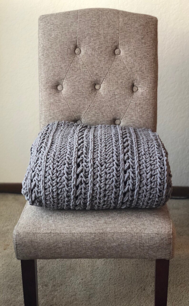 May include: A gray, chunky knit blanket rests on a button-tufted upholstered chair. The chair has a neutral, light brown fabric and dark wooden legs. The blanket's texture is visible, with a thick, woven pattern.