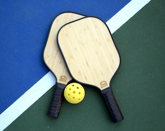 Könnte beinhalten: Zwei hölzerne Pickleball-Schläger mit schwarzem Rand und schwarzen und roten Griffen. Die Schläger liegen auf einem blauen und grünen Platz mit einem gelben Pickleball in der Mitte.