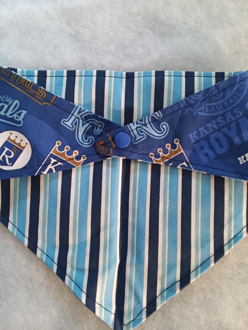 K&ouml;nnte beinhalten: Halsband f&uuml;r Haustiere mit dem Logo des Baseballteams Kansas City Royals. Das Bandana hat blaue und wei&szlig;e Streifen und einen Knopfverschluss. Die obere Schicht zeigt das Teamlogo, die untere Schicht hat vertikale Streifen.