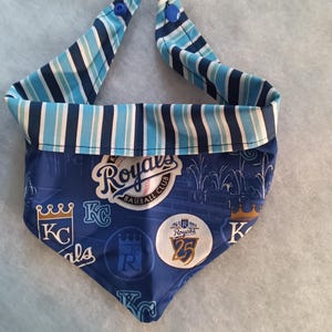 K&ouml;nnte beinhalten: Ein blau-wei&szlig; gestreiftes Hunde-Bandana mit blauem Hintergrund. Das Bandana zeigt das Logo und den Text des Baseballteams Kansas City Royals. Das Bandana hat zwei blaue Druckkn&ouml;pfe.