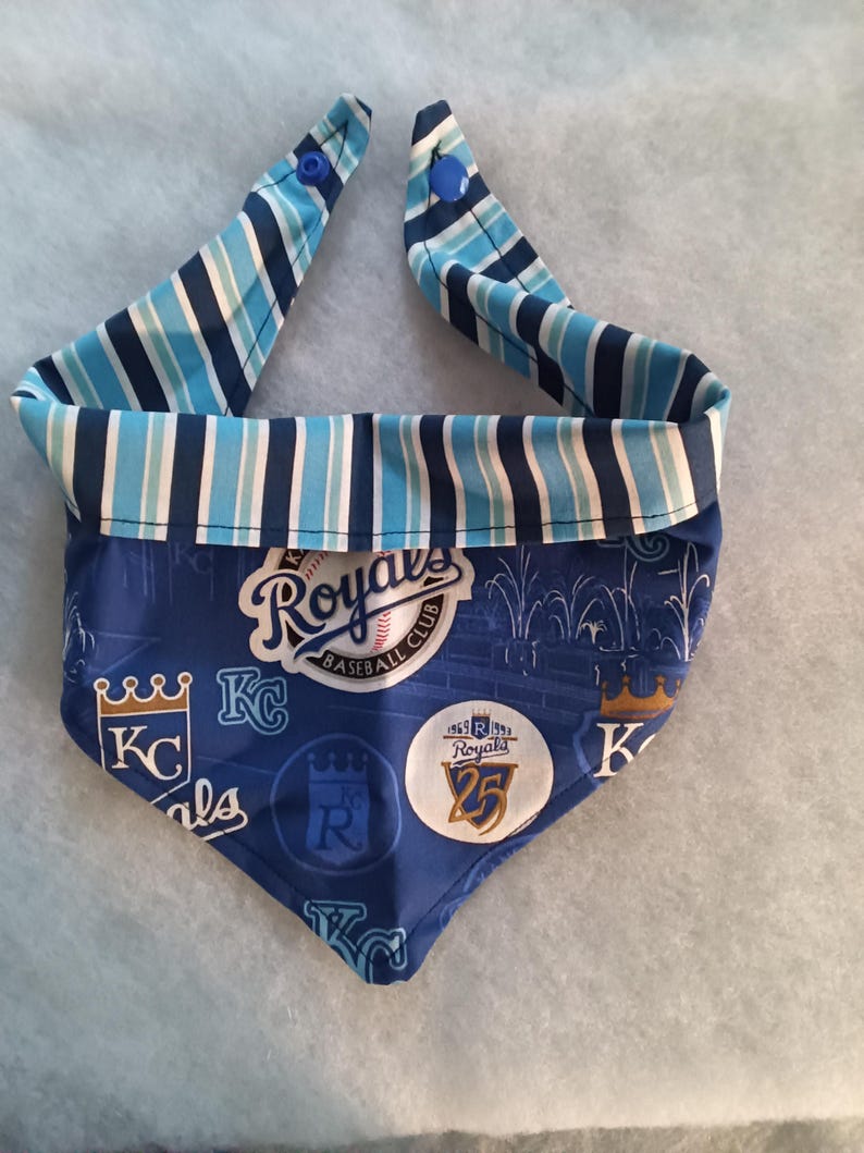 K&ouml;nnte beinhalten: Ein blau-wei&szlig; gestreiftes Hunde-Bandana mit blauem Hintergrund. Das Bandana zeigt das Logo und den Text des Baseballteams Kansas City Royals. Das Bandana hat zwei blaue Druckkn&ouml;pfe.