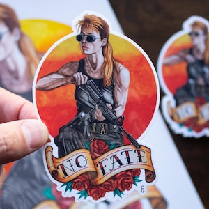 May include: A round sticker featuring a woman with sunglasses holding a rifle, set against a red and yellow background. The sticker has a banner with the words "NO FATE" and red roses. Additional stickers and a print are in the background.