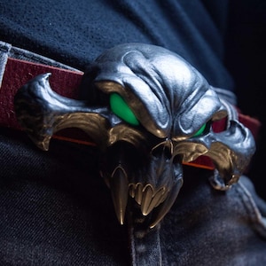 May include: A metallic silver belt buckle shaped like a stylised creature skull with prominent fangs and glowing green eyes. The buckle is attached to a dark red leather belt worn over dark denim jeans, featuring a detailed, textured surface.