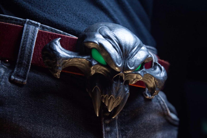 May include: A metallic silver belt buckle shaped like a stylised creature skull with prominent fangs and glowing green eyes. The buckle is attached to a dark red leather belt worn over dark denim jeans, featuring a detailed, textured surface.