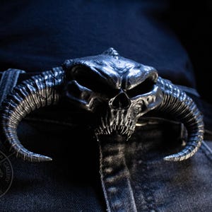 May include: A silver-toned metal belt buckle featuring a detailed, three-dimensional horned skull design with deep eye sockets and sharp teeth. The buckle is attached to a black belt worn over dark denim jeans, showcasing a textured, weathered finish.