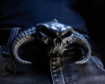 May include: A silver-toned metal belt buckle featuring a detailed, three-dimensional horned skull design with deep eye sockets and sharp teeth. The buckle is attached to a black belt worn over dark denim jeans, showcasing a textured, weathered finish.