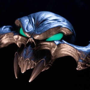 May include: A metallic, dark-toned skull sculpture featuring prominent, elongated fangs and glowing, vibrant green eyes. The skull has a textured, weathered surface with deep contours and wide, sweeping cheekbones that extend outwards to the sides.