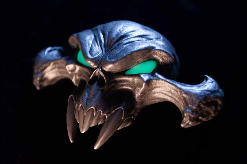 May include: A metallic, dark-toned skull sculpture featuring prominent, elongated fangs and glowing, vibrant green eyes. The skull has a textured, weathered surface with deep contours and wide, sweeping cheekbones that extend outwards to the sides.