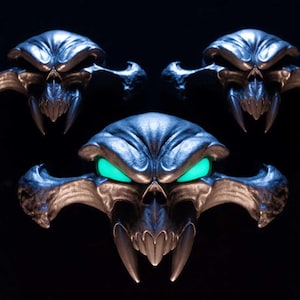 May include: Three metallic skull-shaped ornaments with wide, pointed cheekbones and sharp fangs. The central skull features glowing, bright green eyes, while the two smaller skulls above it have dark, hollow eye sockets, all set against a black background.