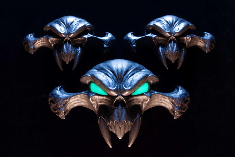 May include: Three metallic skull-shaped ornaments with wide, pointed cheekbones and sharp fangs. The central skull features glowing, bright green eyes, while the two smaller skulls above it have dark, hollow eye sockets, all set against a black background.