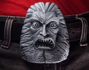 May include: A grey belt buckle featuring a sculpted, three-dimensional face with a wide-eyed, open-mouthed expression and textured, flowing hair. The buckle is attached to a black belt worn over dark denim jeans and a red shirt background.