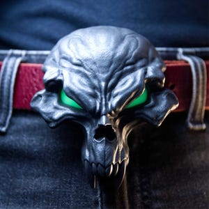 Puede incluir: Una hebilla de cinturón gris oscuro con forma de cráneo estilizado con colmillos prominentes y ojos verdes brillantes. La hebilla está sujeta a un cinturón de cuero rojo sobre pantalones vaqueros oscuros, con una superficie texturizada y contornos.