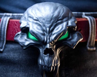 May include: A dark grey belt buckle shaped like a stylised skull with prominent fangs and glowing green eyes. The buckle is attached to a red leather belt worn over dark denim jeans, featuring a textured surface and deep, angular facial contours.