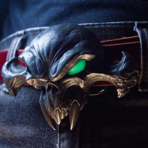 May include: A belt buckle featuring a dark, metallic, stylised skull with prominent fangs and glowing green eyes. The buckle is attached to a red leather belt, which is worn over dark denim jeans. The skull design has a textured, weathered finish.