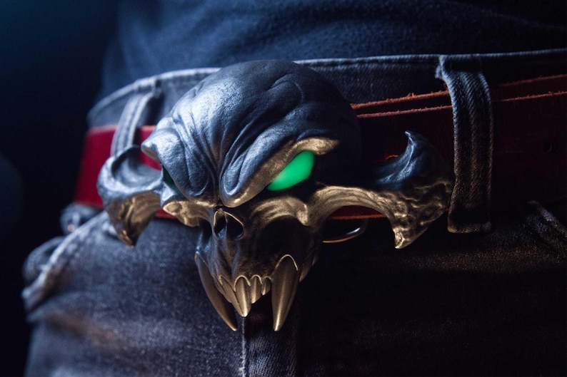 May include: A belt buckle featuring a dark, metallic, stylised skull with prominent fangs and glowing green eyes. The buckle is attached to a red leather belt, which is worn over dark denim jeans. The skull design has a textured, weathered finish.