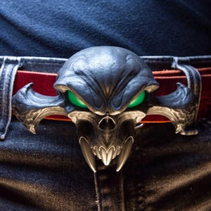 May include: A dark metallic belt buckle shaped like a stylised skull with sharp fangs and glowing green eyes. The buckle is attached to a deep red leather belt, which is threaded through the belt loops of a pair of dark blue denim jeans.