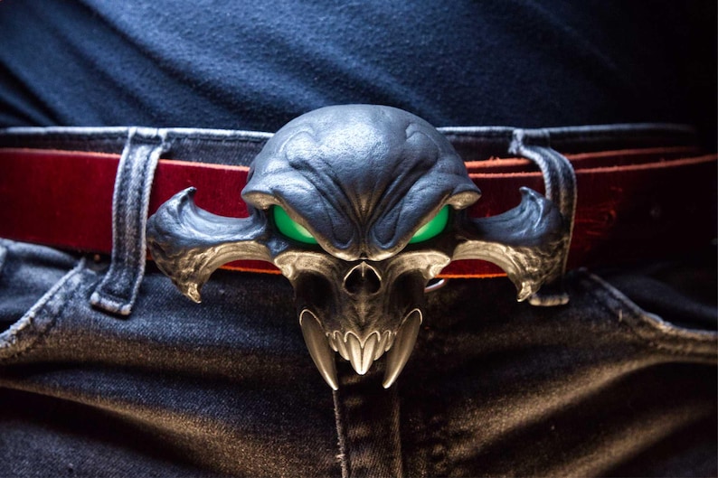 May include: A dark metallic belt buckle shaped like a stylised skull with sharp fangs and glowing green eyes. The buckle is attached to a deep red leather belt, which is threaded through the belt loops of a pair of dark blue denim jeans.