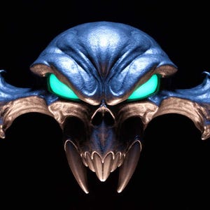 May include: A metallic, dark blue, stylised skull sculpture featuring prominent, sharp fangs and wide, glowing neon green eyes. The object has a smooth, reflective surface with intricate, symmetrical contours and wing-like protrusions on both sides.