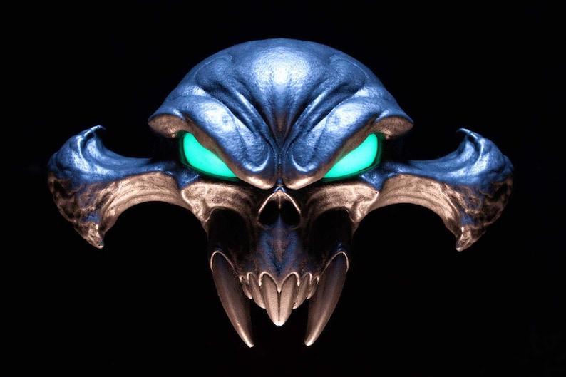 May include: A metallic, dark blue, stylised skull sculpture featuring prominent, sharp fangs and wide, glowing neon green eyes. The object has a smooth, reflective surface with intricate, symmetrical contours and wing-like protrusions on both sides.