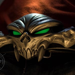 May include: A detailed, metallic skull belt buckle with glowing green eyes. The buckle features sharp teeth and intricate detailing, set against a dark background. The design is reminiscent of a fantasy or comic book theme.