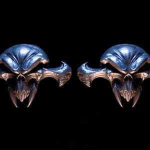 May include: A pair of metallic silver-toned skull-shaped ear gauges featuring a stylised, elongated design with sharp, pointed fangs and wide, sweeping side protrusions. The surface has a polished, reflective finish with deep, dark recessed areas.
