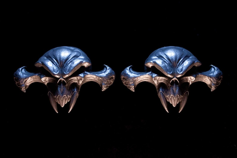 May include: A pair of metallic silver-toned skull-shaped ear gauges featuring a stylised, elongated design with sharp, pointed fangs and wide, sweeping side protrusions. The surface has a polished, reflective finish with deep, dark recessed areas.