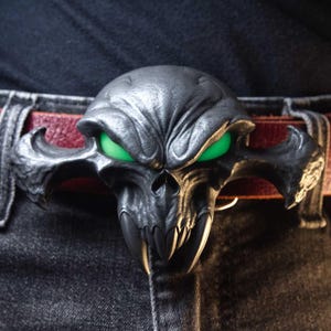 May include: A dark metallic belt buckle shaped like a stylised skull with prominent fangs and glowing green eyes. The buckle is attached to a deep red leather belt worn over dark grey denim jeans, creating a high-contrast accessory piece.