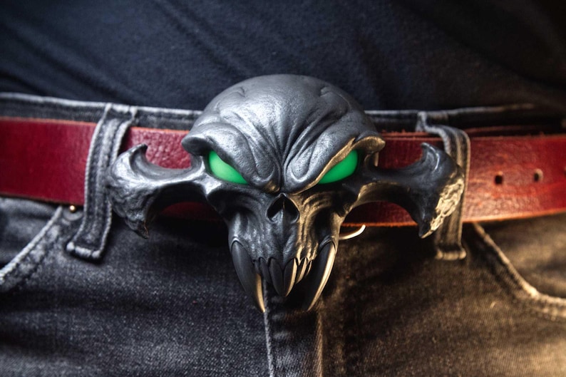 May include: A dark metallic belt buckle shaped like a stylised skull with prominent fangs and glowing green eyes. The buckle is attached to a deep red leather belt worn over dark grey denim jeans, creating a high-contrast accessory piece.