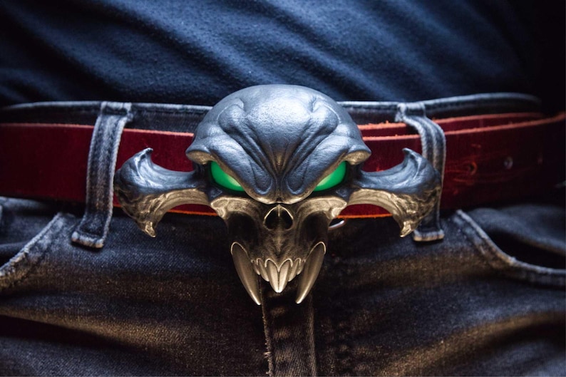 May include: A belt buckle featuring a dark, stylised skull with prominent fangs and glowing green eyes, attached to a deep red leather belt worn over dark denim jeans. The buckle has an intricate, sculpted design with sharp, wing-like side extensions.