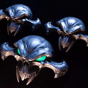 May include: Three dark metallic skull-shaped ornaments with wide, pointed cheekbones and sharp fangs. The largest skull in the foreground features glowing green eyes, while the two smaller skulls above it have dark, hollow eye sockets.