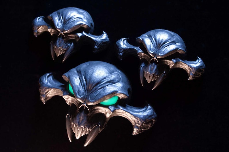 May include: Three dark metallic skull-shaped ornaments with wide, pointed cheekbones and sharp fangs. The largest skull in the foreground features glowing green eyes, while the two smaller skulls above it have dark, hollow eye sockets.