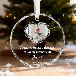 May include: A clear, faceted glass ornament with a photo of a man inside a heart-shaped frame. The ornament has the text "Forever in our hearts, In Loving Memory Of Jovial 1961-2025". A white ribbon is attached for hanging.