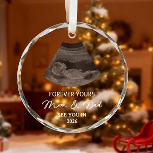Op de afbeelding: Een heldere, ronde kerstversiering met een lint om op te hangen. De versiering toont een echografiebeeld en de tekst "FOREVER YOURS Mom & Dad SEE YOU IN 2026". De achtergrond omvat een kerstboom en een rode slee.