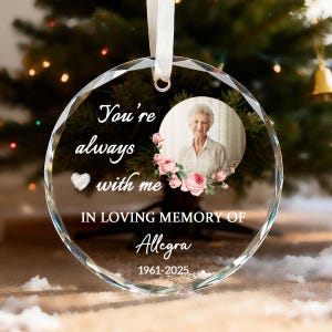 May include: A clear, round crystal ornament with a photo of a woman surrounded by pink roses. Text reads "You're always with me" and "In Loving Memory of Allegra 1961-2025". A white ribbon is attached for hanging.