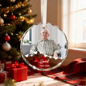 May include: A clear, round Christmas ornament with a photo of a man in a striped shirt. The ornament has a white ribbon for hanging and the name "David" with the years 1955-2025. The background includes a Christmas tree and wrapped gifts.