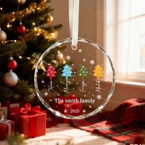 May include: A clear, circular Christmas bauble with a white ribbon. It displays four colourful Christmas trees and names, with the text "The smith family 2025". A Christmas tree and wrapped gifts are in the background.