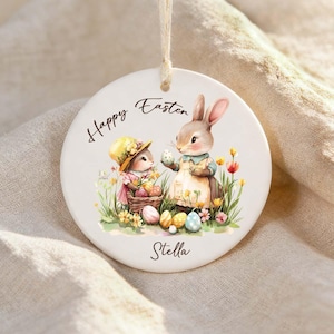 May include: Round ceramic Easter ornament with a watercolour illustration of two bunnies, Easter eggs, and flowers. The ornament has the words "Happy Easter" and the name "Stella" printed on it. A string is attached for hanging.