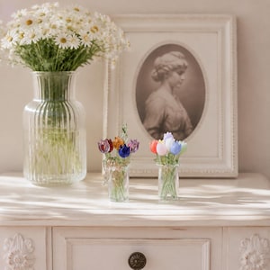 Puede incluir: Un jarrón grande de cristal transparente acanalado lleno de margaritas blancas, junto a dos jarrones pequeños de cristal transparente con tulipanes de cristal en miniatura de varios colores, incluyendo rojo, naranja, azul, morado, rosa y verde claro.