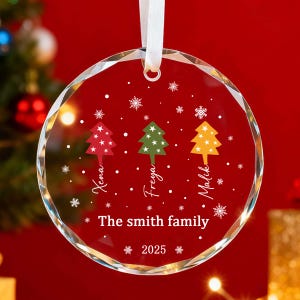 May include: A clear, circular Christmas bauble with a red backdrop. It displays three Christmas trees in red, green, and yellow, with names beneath each. The text "The smith family" and "2025" are also present. A white ribbon is attached.