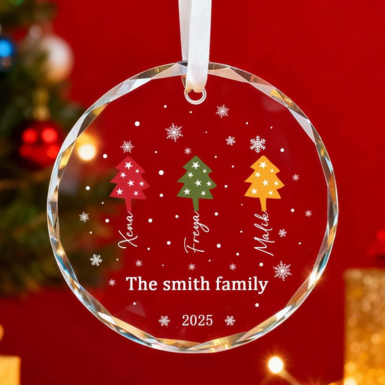 May include: A clear, circular Christmas bauble with a red backdrop. It displays three Christmas trees in red, green, and yellow, with names beneath each. The text "The smith family" and "2025" are also present. A white ribbon is attached.