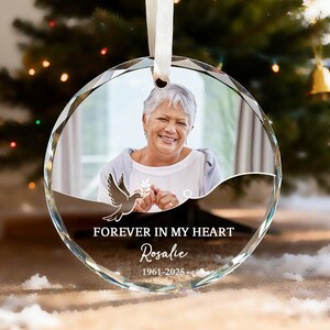 May include: A clear, round crystal ornament with a photo of a smiling woman. The ornament has a white ribbon for hanging and features the text "FOREVER IN MY HEART" and the name "Rosalie" with the years 1961-2025. A dove graphic is also present.